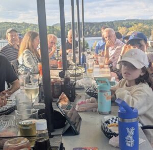 A group of people of all ages site around a table overlooking a lake.