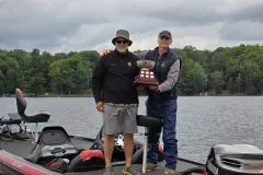 Lou and Joe stand in a boat holding the Percy Lake bass tournament trophy.