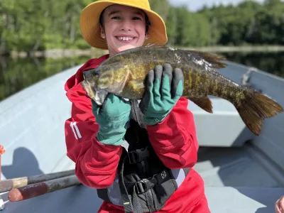 A young Arlo wearing gloves holds up a bass while sitting in a boat.