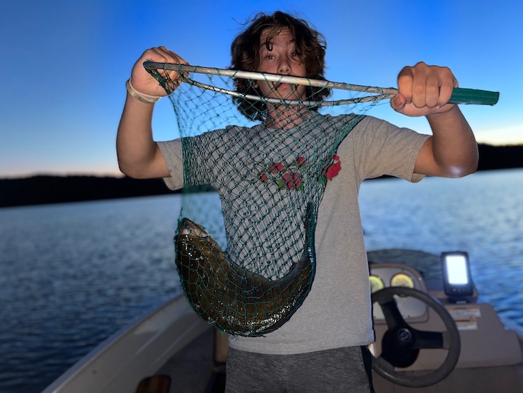 Julian Tilman stands on a boat in the middle of a lake, holding a fishing net with a bass inside.